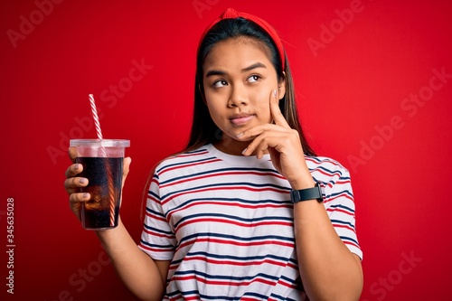 Tableau sur toile Young asian girl drinking cola fizzy refreshment using straw over isolated red b
