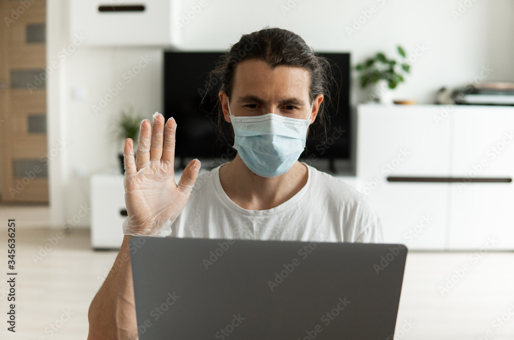 Man in medical mask is working from home with a laptop and showing hello to his friends or business partners via webcam because of coronavirus epidemic. Remote work during pandemic COVID-19. Man in medical mask is working from home with a laptop and showing hello to his friends or business partners via webcam because of coronavirus epidemic. Remote work during pandemic COVID-19.