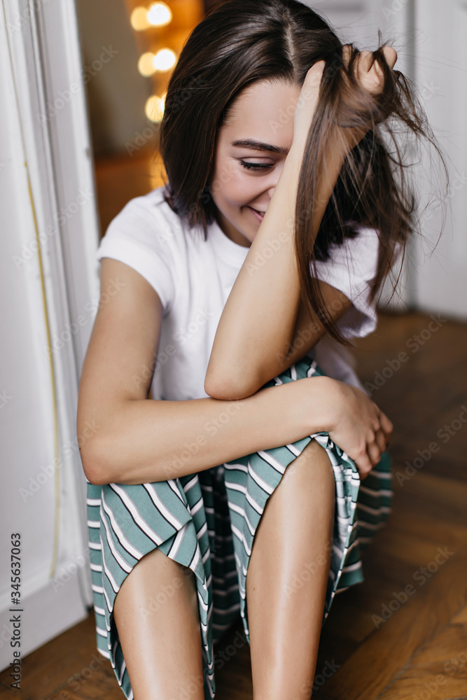 Fototapeta premium Opalona dziewczyna w ślicznej piżamie śmiejąca się i bawiąca się włosami. Wewnątrz zdjęcie wyrafinowanej młodej damy siedzącej na podłodze ze szczerym uśmiechem.