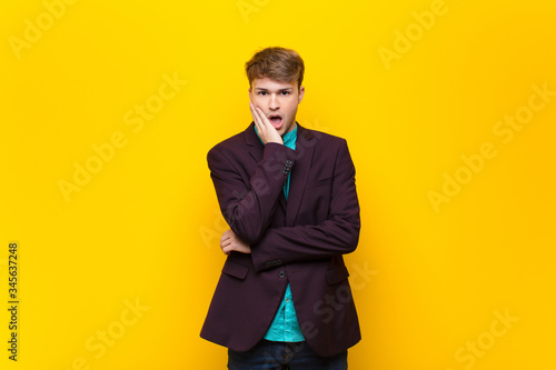 young blonde man open-mouthed in shock and disbelief, with hand on cheek and arm crossed, feeling stupefied and amazed isolated against flat wall photo