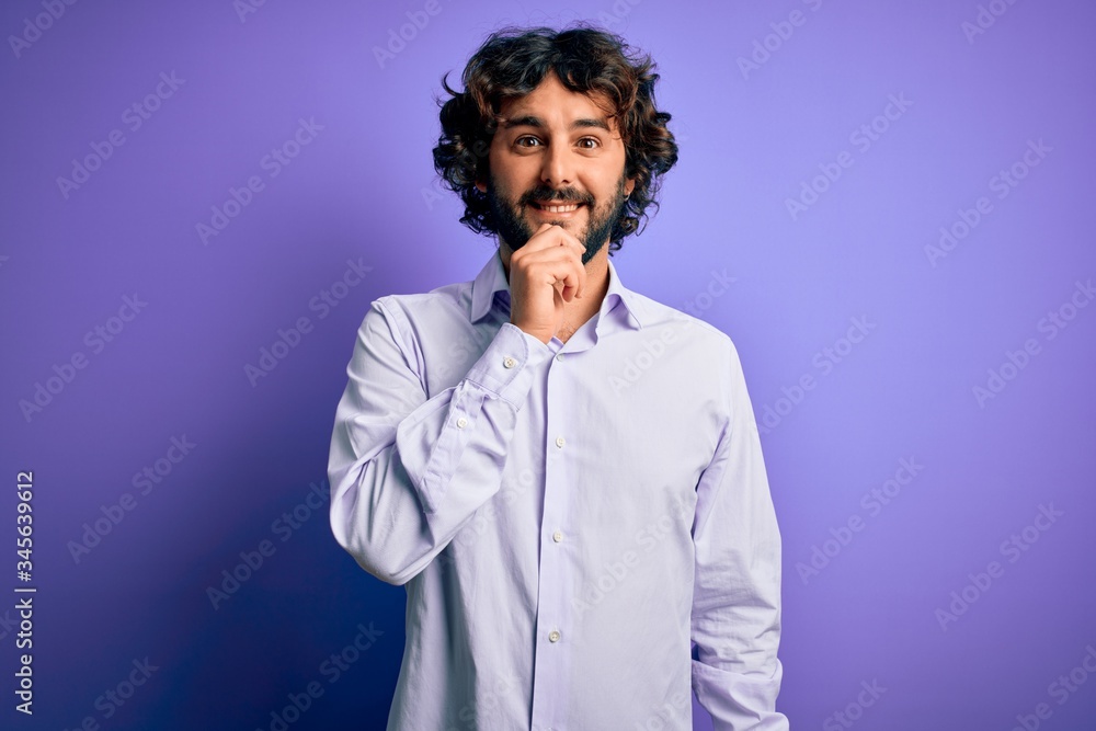 Young handsome business man with beard wearing shirt standing over purple background looking confident at the camera smiling with crossed arms and hand raised on chin. Thinking positive.