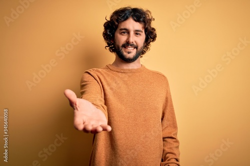 Fotografie Young handsome man with beard wearing casual sweater standing over yellow background smiling cheerful offering palm hand giving assistance and acceptance
