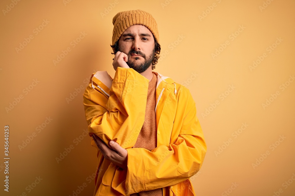 Young handsome man with beard wearing raincoat for rainy day over yellow background thinking looking tired and bored with depression problems with crossed arms.