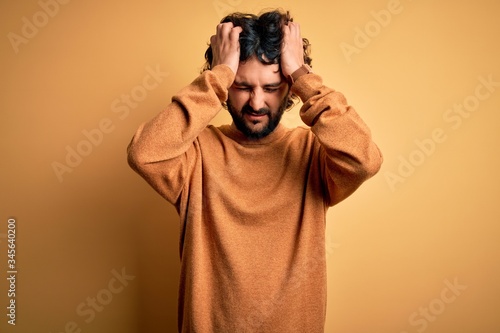 Young handsome man with beard wearing casual sweater standing over yellow background suffering from headache desperate and stressed because pain and migraine. Hands on head.