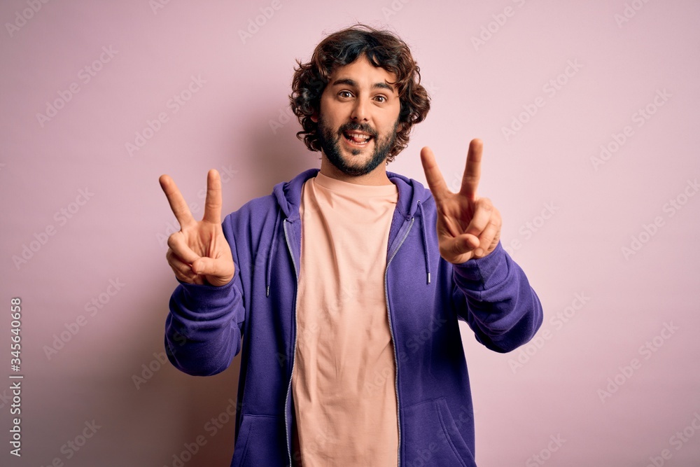 Young handsome sporty man with beard wearing casual sweatshirt over pink background smiling with tongue out showing fingers of both hands doing victory sign. Number two. Young handsome sporty man with beard wearing casual sweatshirt over pink background smiling with tongue out showing fingers of both hands doing victory sign. Number two.