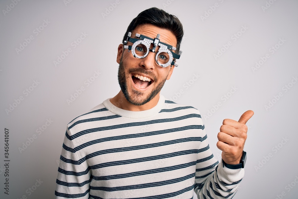 Young handsome man with beard wearing optometry glasses over isolated white background smiling with happy face looking and pointing to the side with thumb up. Young handsome man with beard wearing optometry glasses over isolated white background smiling with happy face looking and pointing to the side with thumb up.