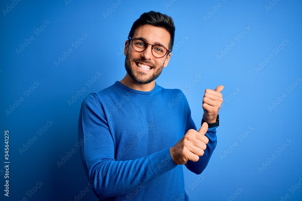 Young handsome man with beard wearing casual sweater and glasses over blue background Pointing to the back behind with hand and thumbs up, smiling confident Young handsome man with beard wearing casual sweater and glasses over blue background Pointing to the back behind with hand and thumbs up, smiling confident