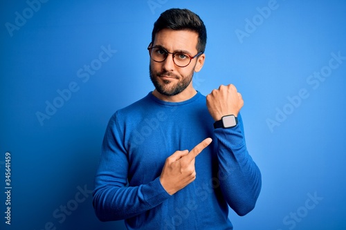Young handsome man with beard wearing casual sweater and glasses over blue background In hurry pointing to watch time, impatience, looking at the camera with relaxed expression