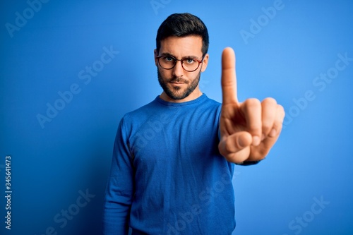 Young handsome man with beard wearing casual sweater and glasses over blue background Pointing with finger up and angry expression, showing no gesture photo
