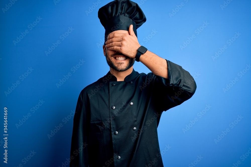 Young handsome chef man with beard wearing cooker uniform and hat over blue background smiling and laughing with hand on face covering eyes for surprise. Blind concept. Young handsome chef man with beard wearing cooker uniform and hat over blue background smiling and laughing with hand on face covering eyes for surprise. Blind concept.
