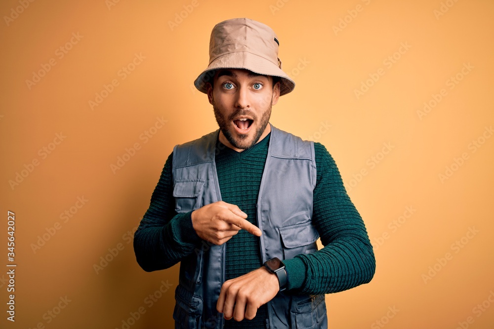 Handsome tourist man with beard on vacation wearing explorer hat over yellow background In hurry pointing to watch time, impatience, upset and angry for deadline delay Handsome tourist man with beard on vacation wearing explorer hat over yellow background In hurry pointing to watch time, impatience, upset and angry for deadline delay