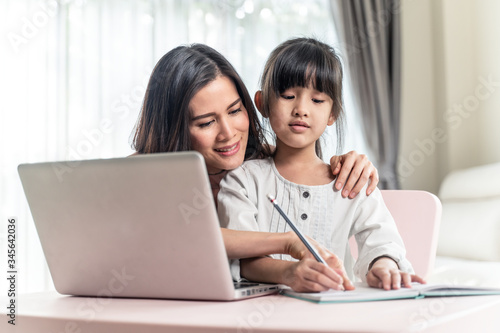 Homeschool Asian young little girl learning online and does homework by using internet on computer with mother help, teach and encourage. Daughter smile and happy to study at home together with mom.