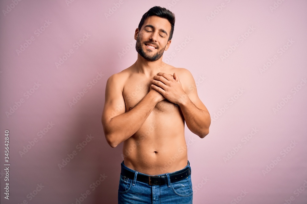 Young handsome strong man with beard shirtless standing over isolated pink background smiling with hands on chest with closed eyes and grateful gesture on face. Health concept.