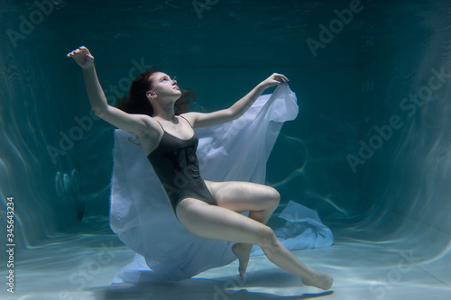 Fototapeta Naklejka Na Ścianę i Meble -  Beautiful girl swims underwater with long hair. Blue or gold background like gold. The atmosphere of a fairy tale or magic. Diving under the water with a shiny cloth