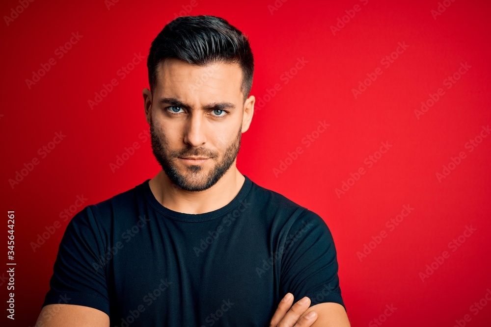 Young handsome man wearing casual black t-shirt standing over isolated red background skeptic and nervous, disapproving expression on face with crossed arms. Negative person. Young handsome man wearing casual black t-shirt standing over isolated red background skeptic and nervous, disapproving expression on face with crossed arms. Negative person.