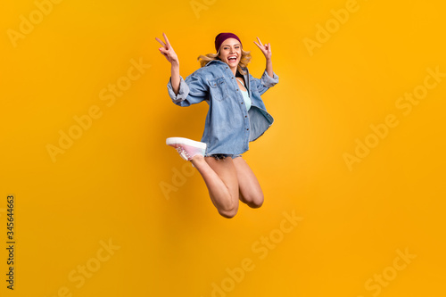 Full length photo of attractive lady long hairdo good mood students party jump high up showing v-sign symbols wear denim shirt shorts hat shoes isolated yellow color background