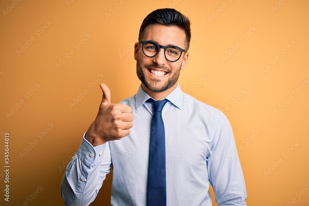 Young handsome businessman wearing tie and glasses standing over yellow background doing happy thumbs up gesture with hand. Approving expression looking at the camera showing success. Young handsome businessman wearing tie and glasses standing over yellow background doing happy thumbs up gesture with hand. Approving expression looking at the camera showing success.