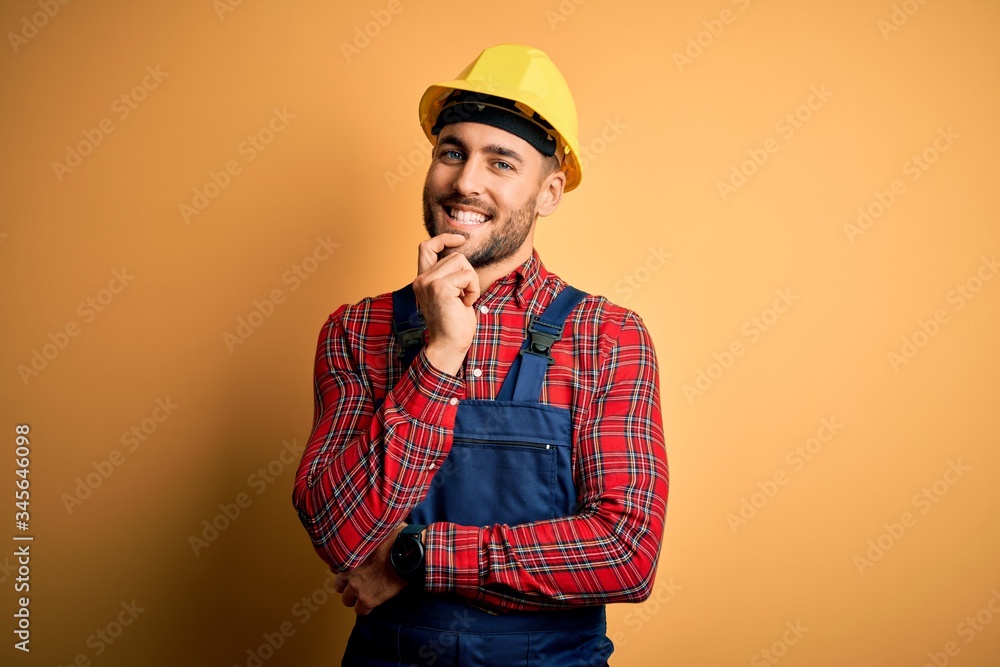 Young builder man wearing construction uniform and safety helmet over yellow isolated background looking confident at the camera with smile with crossed arms and hand raised on chin. Thinking positive