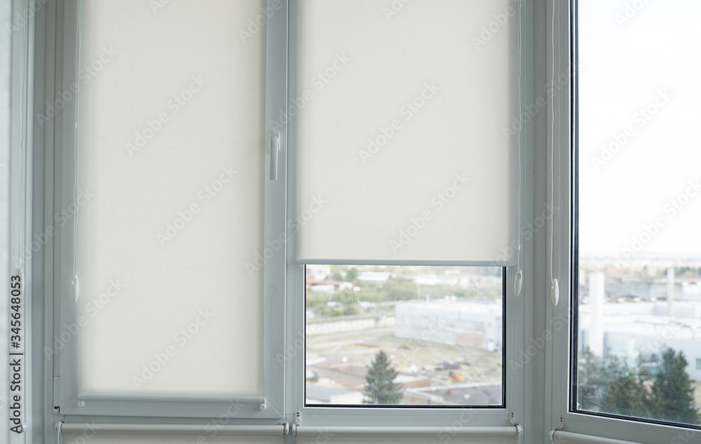 Details of white fabric roller blinds on the plastic window with wood texture in the living room. Details of white fabric roller blinds on the plastic window with wood texture in the living room.