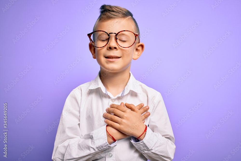 Young little caucasian kid with blue eyes wearing glasses and white shirt over purple background smiling with hands on chest with closed eyes and grateful gesture on face. Health concept. Young little caucasian kid with blue eyes wearing glasses and white shirt over purple background smiling with hands on chest with closed eyes and grateful gesture on face. Health concept.