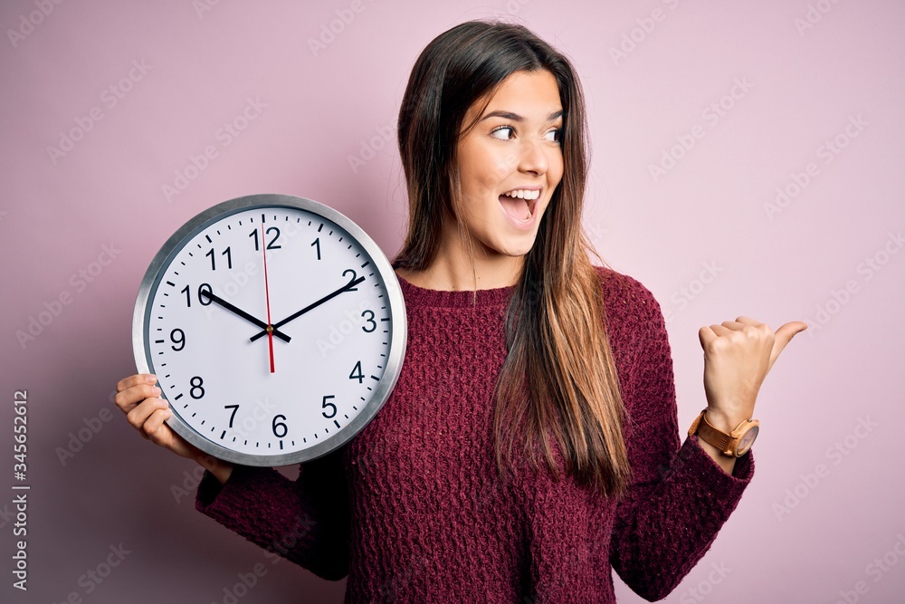 Young beautiful girl doing countdown holding big clock over isolated pink background pointing and showing with thumb up to the side with happy face smiling Young beautiful girl doing countdown holding big clock over isolated pink background pointing and showing with thumb up to the side with happy face smiling