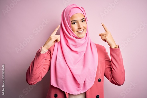 Young beautiful girl wearing muslim hijab standing over isolated pink background smiling pointing to head with both hands finger, great idea or thought, good memory © Krakenimages.com