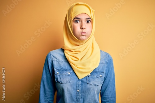 Young beautiful girl wearing muslim hijab standing over isolated yellow background puffing cheeks with funny face. Mouth inflated with air, crazy expression. © Krakenimages.com