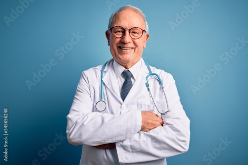 Senior grey haired doctor man wearing stethoscope and medical coat over blue background happy face smiling with crossed arms looking at the camera. Positive person.