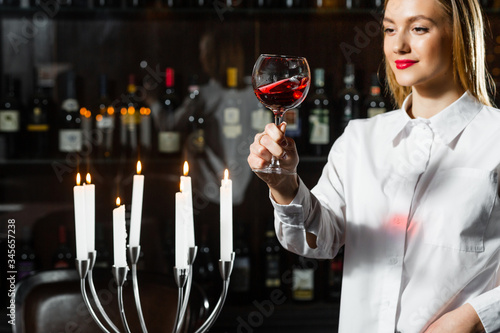 Beautiful blonde young girl sommelier holding up and looking through a glass of wine. Color and bouquet of wine.