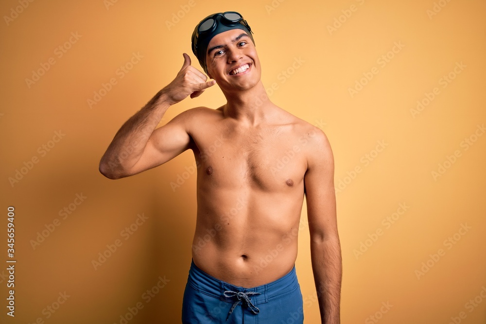 Young handsome man shirtless wearing swimsuit and swim cap over isolated yellow background smiling doing phone gesture with hand and fingers like talking on the telephone. Communicating concepts.