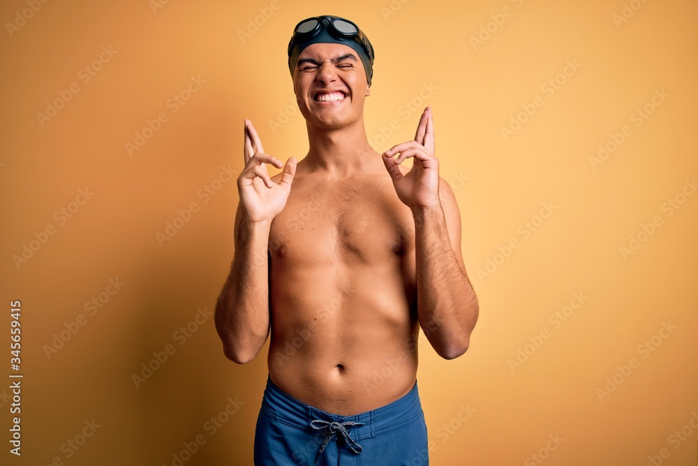 Young handsome man shirtless wearing swimsuit and swim cap over isolated yellow background gesturing finger crossed smiling with hope and eyes closed. Luck and superstitious concept.