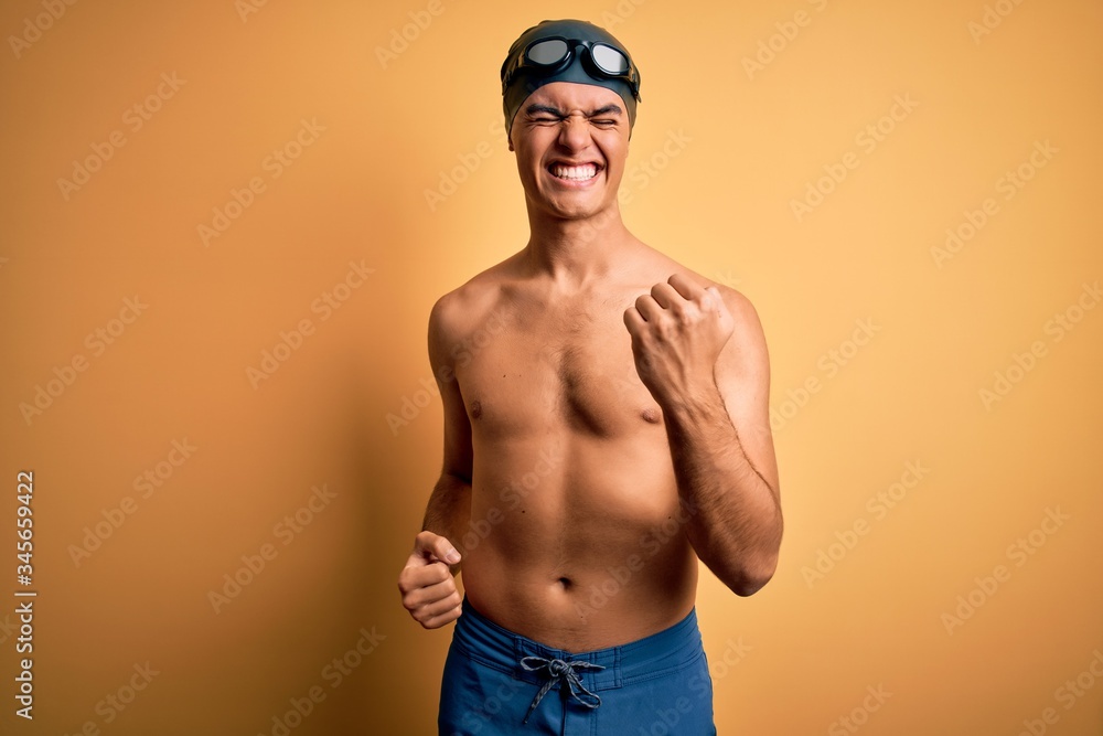 Young handsome man shirtless wearing swimsuit and swim cap over isolated yellow background celebrating surprised and amazed for success with arms raised and eyes closed. Winner concept.