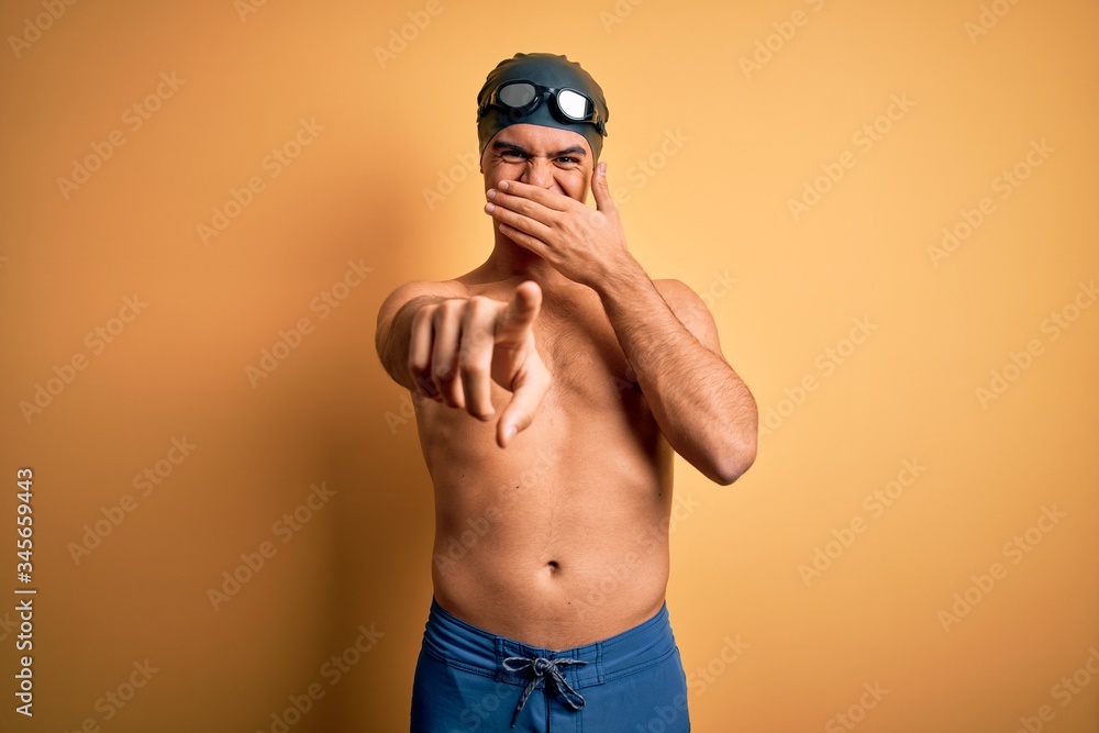 Young handsome man shirtless wearing swimsuit and swim cap over isolated yellow background laughing at you, pointing finger to the camera with hand over mouth, shame expression