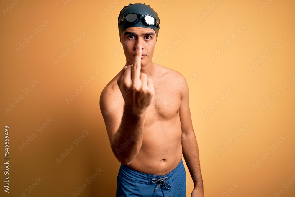 Young handsome man shirtless wearing swimsuit and swim cap over isolated yellow background Showing middle finger, impolite and rude fuck off expression