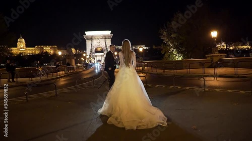 the bride goes to meet the groom against the backdrop of an ancient bridge, old night city, lanterns and light. Budapest. 
meeting young beautiful fashion wedding couple. white dress and black suit.