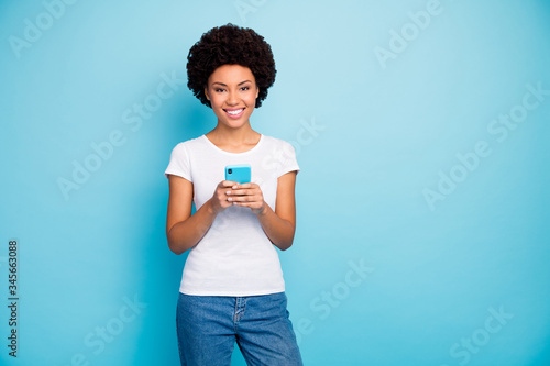 Photo of cheerful beautiful dark skin curly lady holding telephone hands chatting friends sending new pictures wear casual white t-shirt jeans isolated blue color background