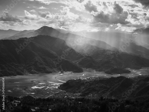 A black and white landscape of sun rays streaming through dramatic clouds to hit the mountains and a valley in Pokhara, Nepal.