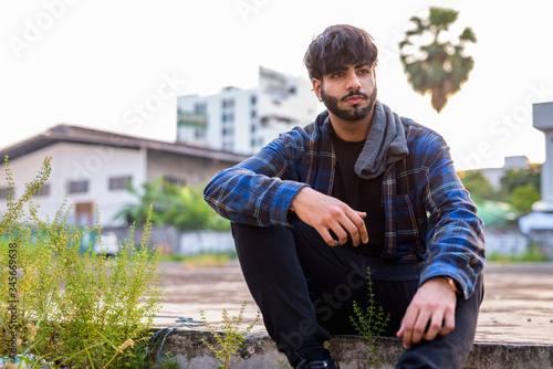 Young handsome bearded Indian hipster man sitting in urban streets