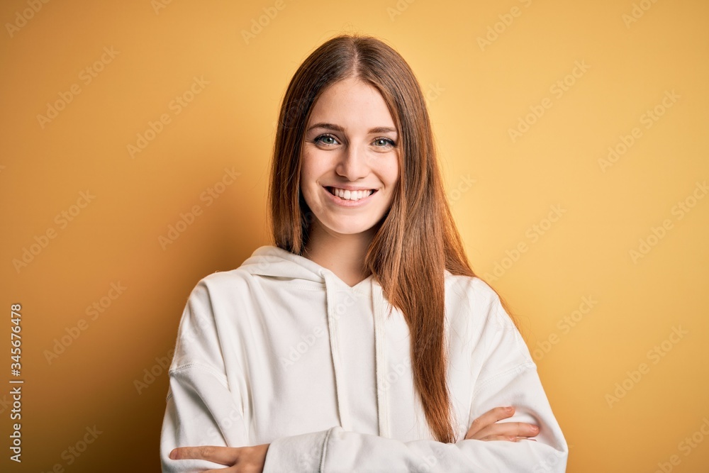 Young beautiful redhead sporty woman wearing sweatshirt over isolated yellow background happy face smiling with crossed arms looking at the camera. Positive person. Young beautiful redhead sporty woman wearing sweatshirt over isolated yellow background happy face smiling with crossed arms looking at the camera. Positive person.