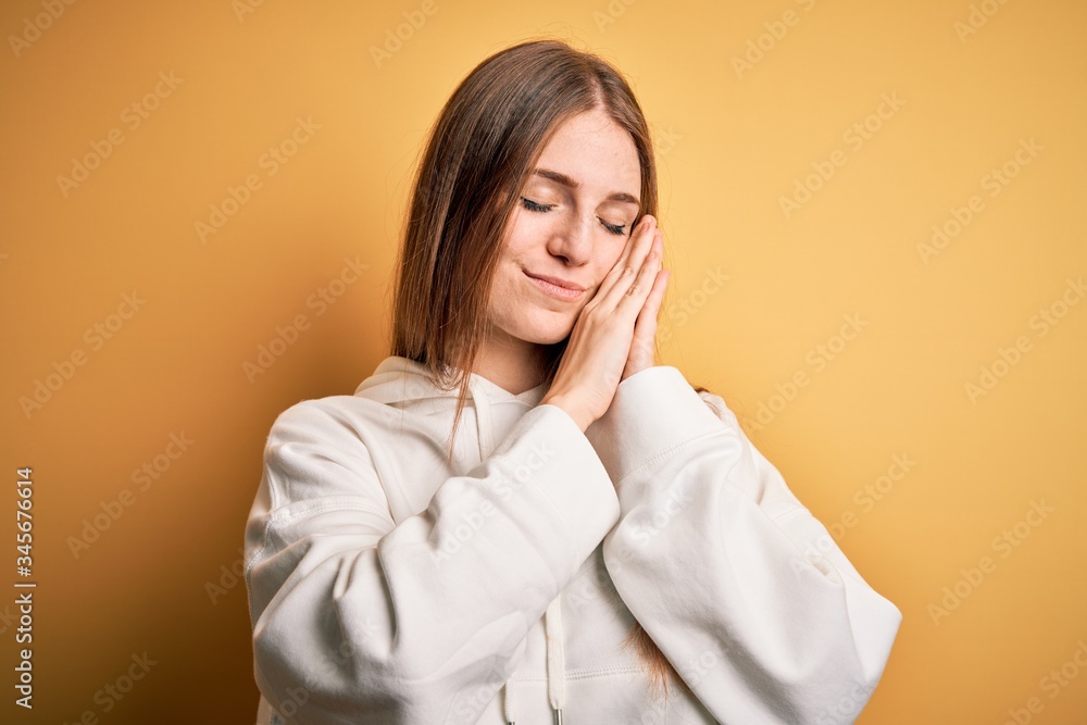 Young beautiful redhead sporty woman wearing sweatshirt over isolated yellow background sleeping tired dreaming and posing with hands together while smiling with closed eyes. Young beautiful redhead sporty woman wearing sweatshirt over isolated yellow background sleeping tired dreaming and posing with hands together while smiling with closed eyes.