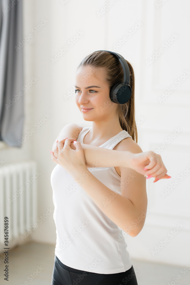A girl of European appearance smiles, stands in black headphones and does shoulder stretching exercises in a bright studio A girl of European appearance smiles, stands in black headphones and does shoulder stretching exercises in a bright studio