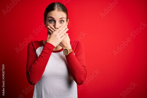 Young beautiful redhead woman wearing casual t-shirt over isolated red background shocked covering mouth with hands for mistake. Secret concept.