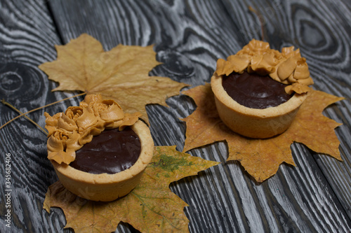 Tartlets with chocolate ganache. On autumn maple leaves. Decorated with oil cream flowers. The cream has a caramel color. On brushed pine boards painted black.