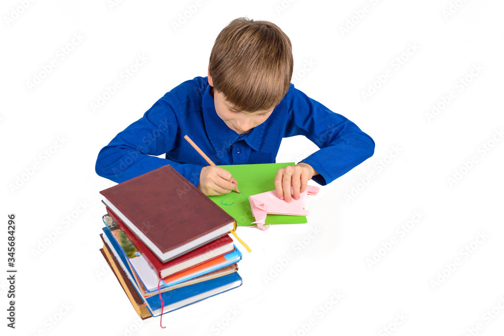 child development: a schoolboy boy in a blue T-shirt sits at the table and draws on colored paper, next to a stack of textbooks and paperwork, origami child development: a schoolboy boy in a blue T-shirt sits at the table and draws on colored paper, next to a stack of textbooks and paperwork, origami