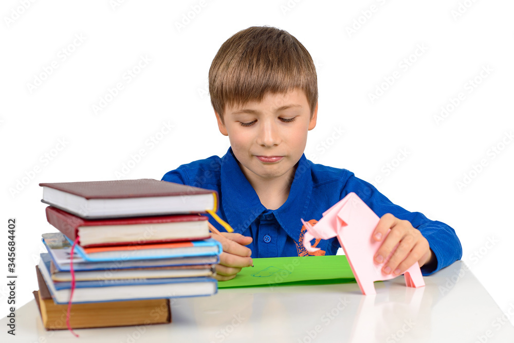 child development: a schoolboy boy in a blue T-shirt sits at the table and draws on colored paper, next to a stack of textbooks and paperwork, origami child development: a schoolboy boy in a blue T-shirt sits at the table and draws on colored paper, next to a stack of textbooks and paperwork, origami