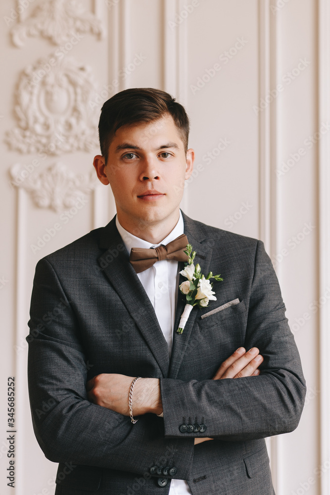 Portrait of the groom. Business young man, in a classic suit, gray with bow-tie and boutonniere. In a classic bright interior. Portrait of the groom. Business young man, in a classic suit, gray with bow-tie and boutonniere. In a classic bright interior.