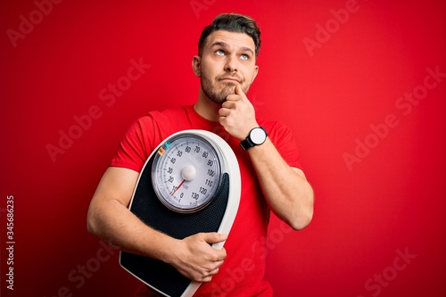 Young fitness man with blue eyes holding scale dieting for healthy weight over red background serious face thinking about question, very confused idea