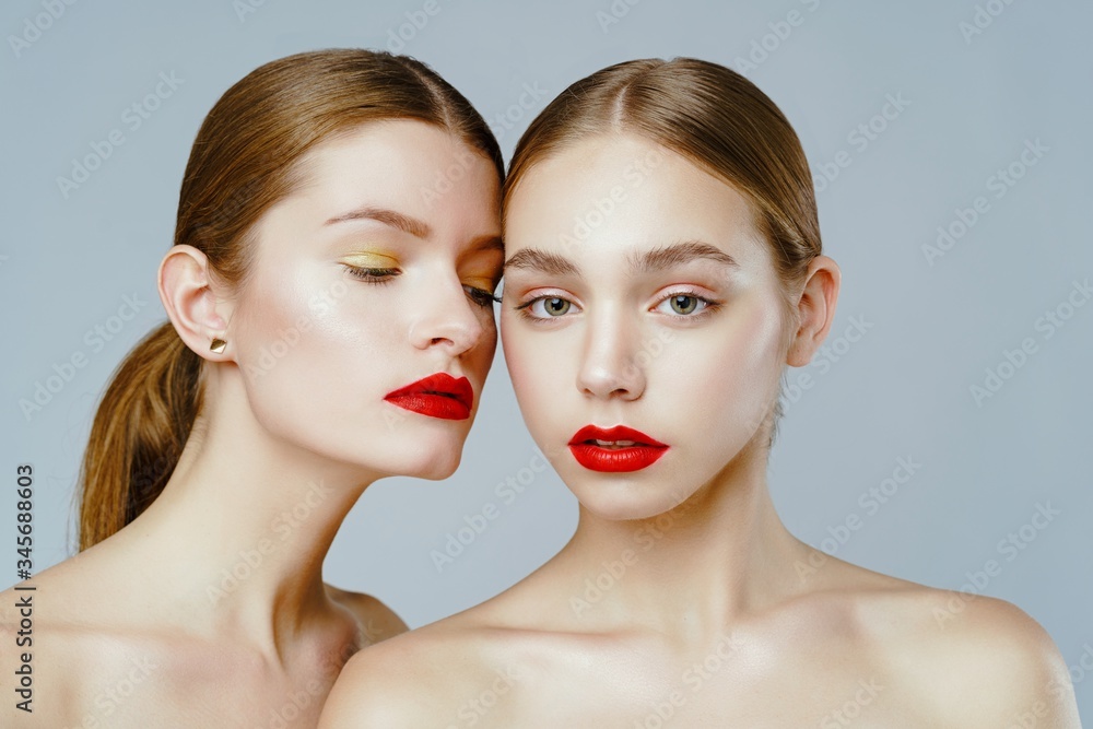 Close-up portrait of two gorgeous young fashion models with red lips posing grey background. Beautiful healthy face of the young pretty woman with fresh skin.