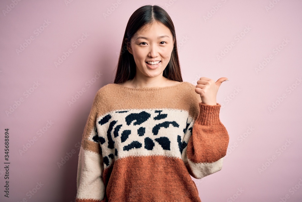 Young beautiful asian woman wearing animal print fashion sweater over pink isolated background smiling with happy face looking and pointing to the side with thumb up. Young beautiful asian woman wearing animal print fashion sweater over pink isolated background smiling with happy face looking and pointing to the side with thumb up.