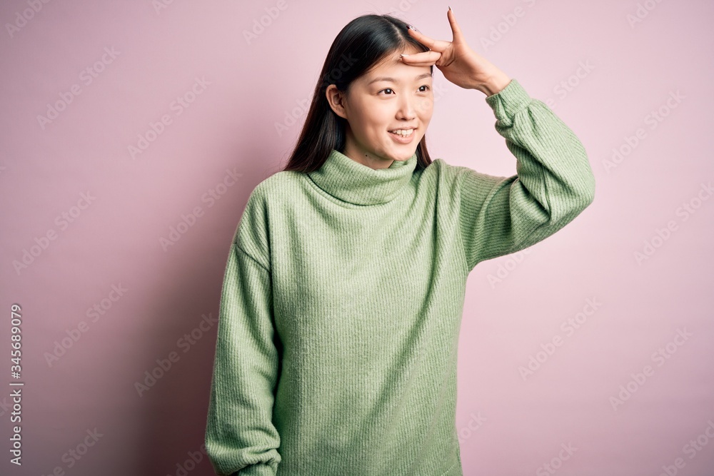Young beautiful asian woman wearing green winter sweater over pink solated background very happy and smiling looking far away with hand over head. Searching concept. Young beautiful asian woman wearing green winter sweater over pink solated background very happy and smiling looking far away with hand over head. Searching concept.